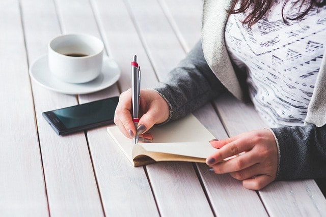 A woman writing in a notepad with her smartphone and a cup of coffee next to her.