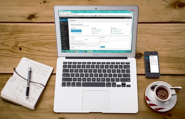 A laptop, a notebook, a smartphone, and a cup of coffee on top of a wooden desk.