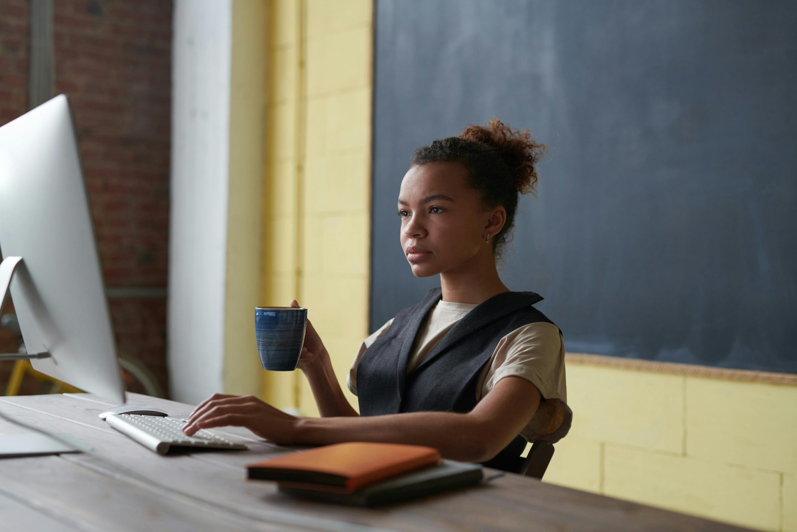 A female teacher sitting at her desk in front of a desktop computer and holding a cup of coffee.