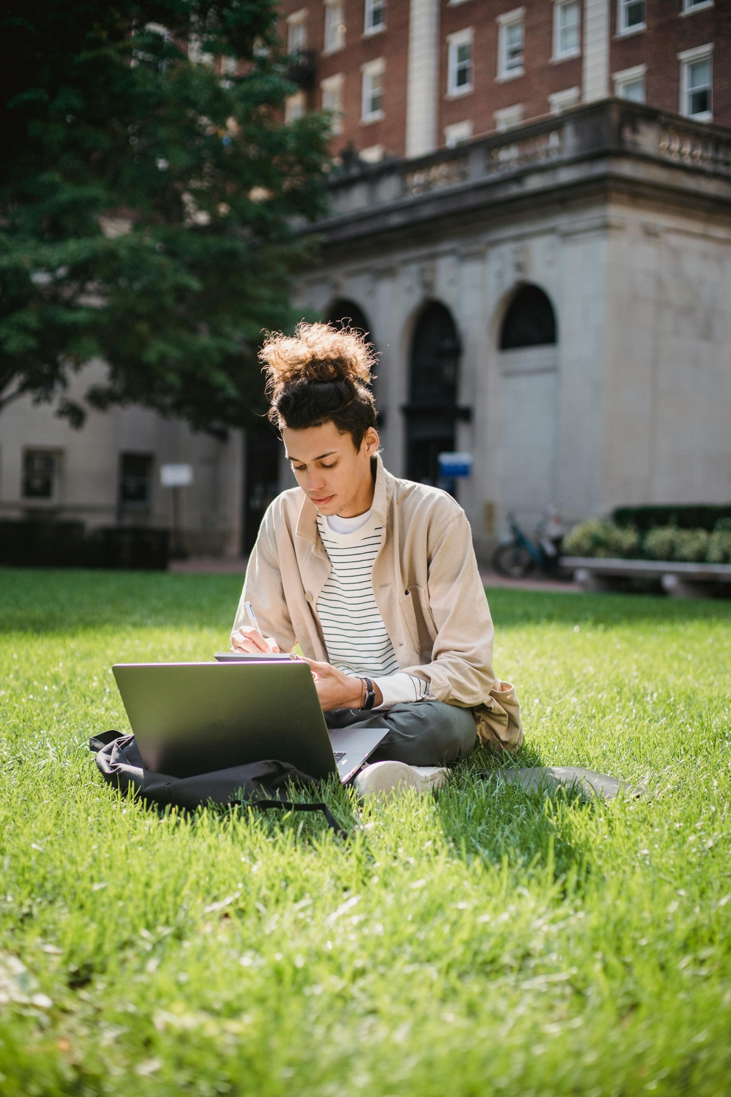 A person sitting on the grass at a university campus and working on their laptop. 