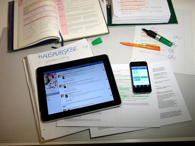 A tablet, a smartphone, a pen, a highlighter, and lots of open notebooks on a desk.