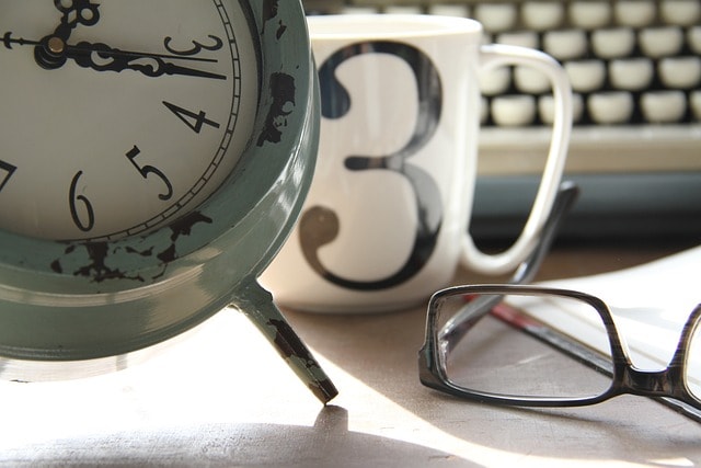 A close-up of a clock, a coffee mug with the number 3 on it, a pair of reading glasses, and a keyboard in the background.