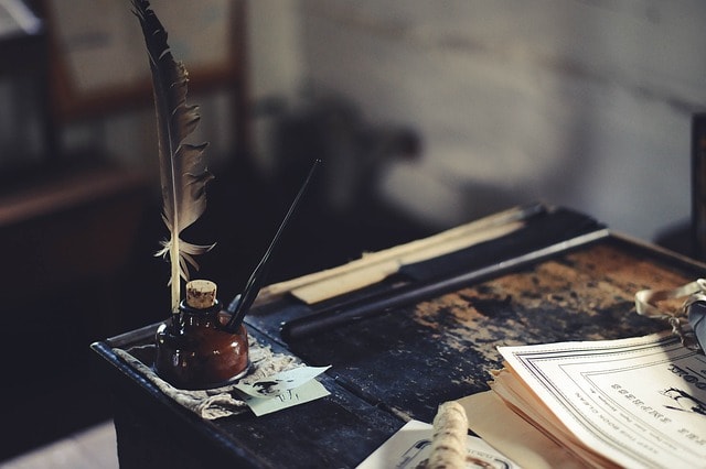 A wooden desk with a quill, an ink jar, and some pieces of paper on top of it.