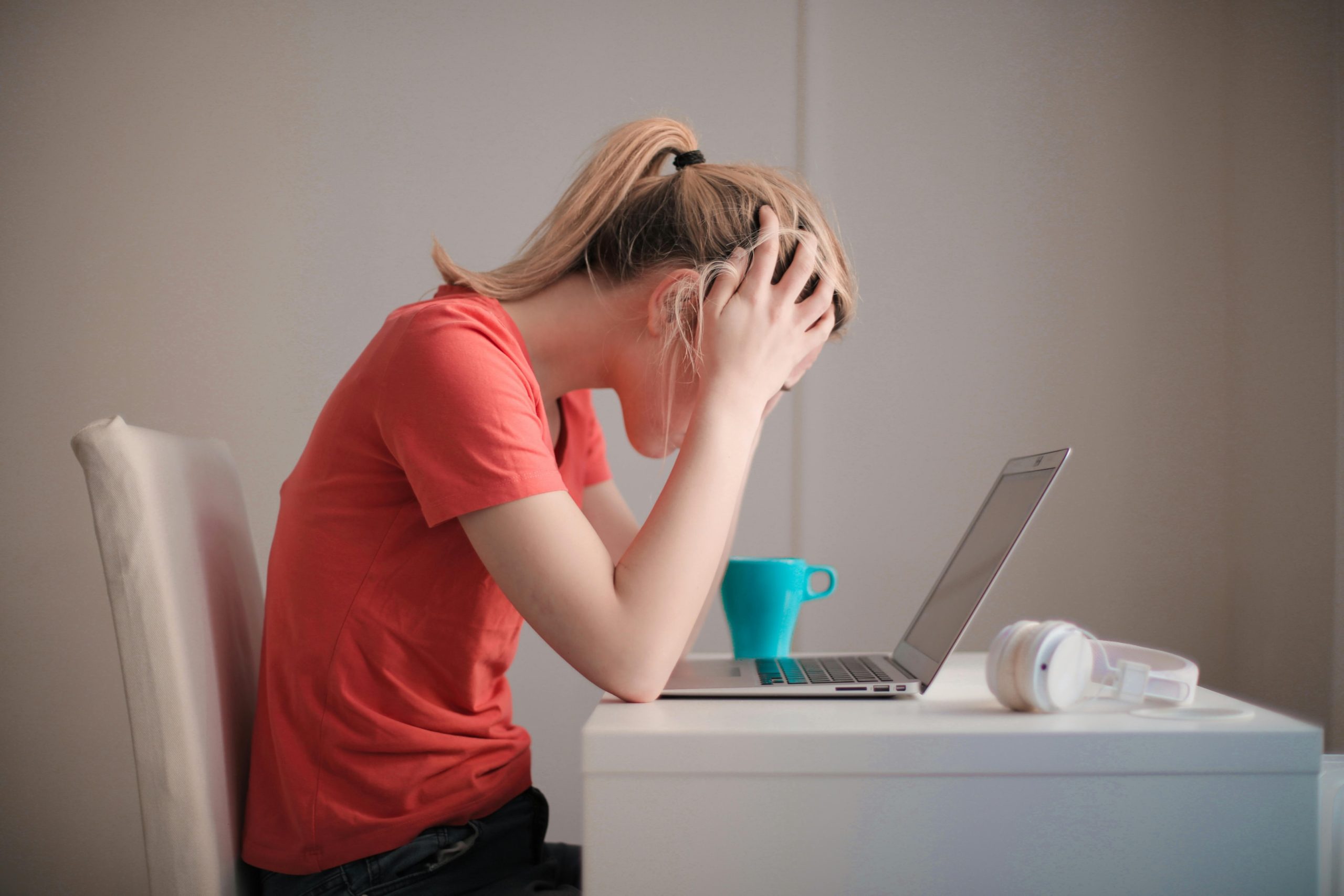  A woman in a red t-shirt holds her head in her hands while sitting in front of her laptop.