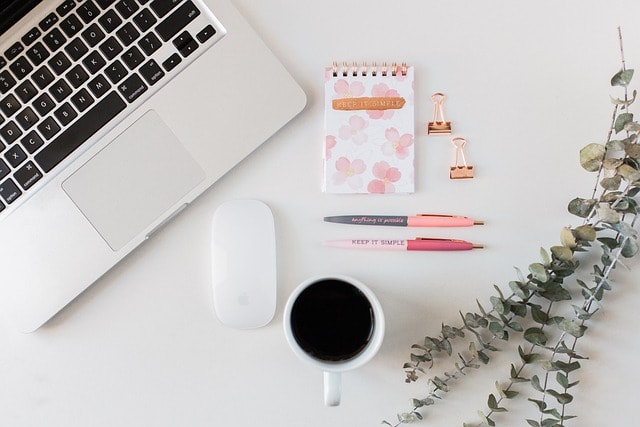 A laptop next to a mouse, a cup of coffee, 2 pink pens, and a notebook.