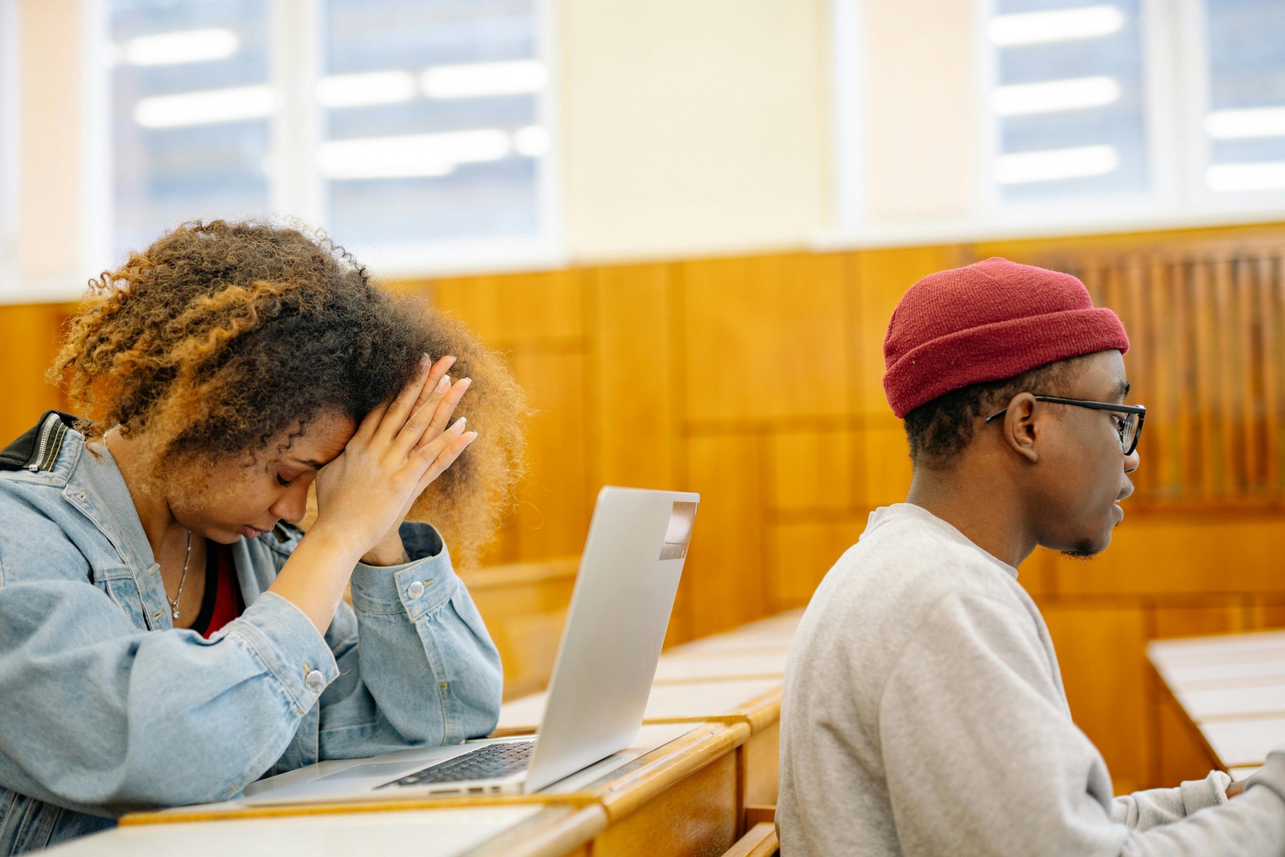 Studente, kas sēž aiz studenta vīrieša eksāmenu zālē, sadevusi rokas kopā.