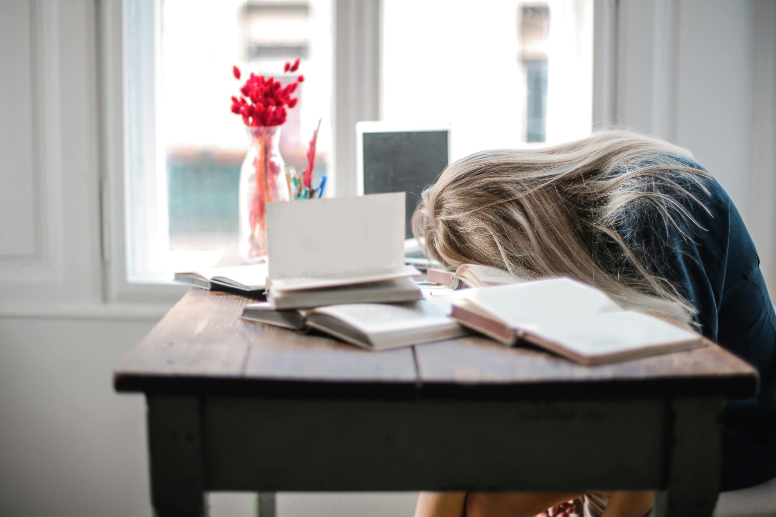 A tired woman resting her head on her work desk while surrounded by books.