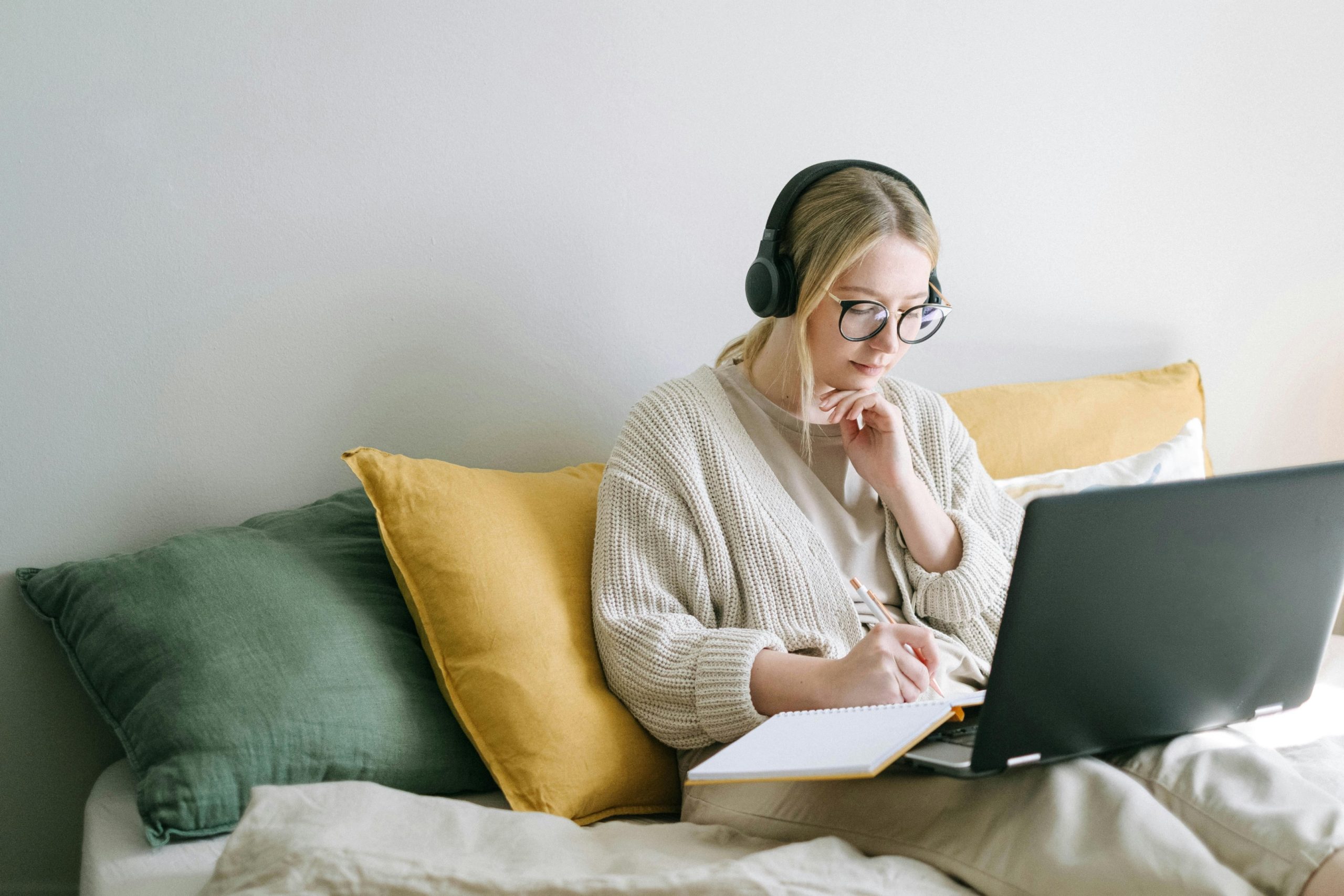 A woman taking notes in a notebook while sitting on a sofa with headphones on. 