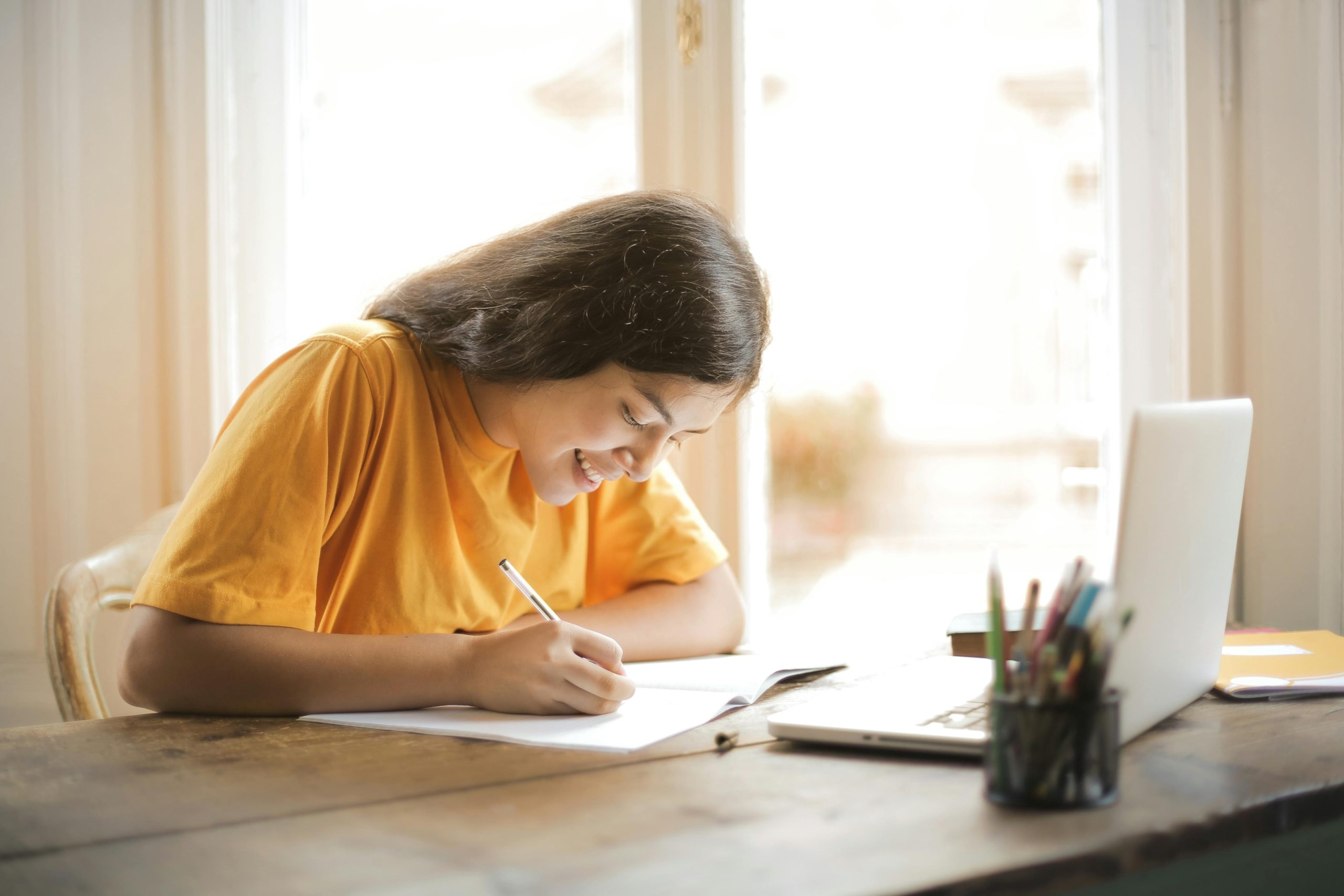 A girl in a yellow t-shirt working on a desk in front of a laptop and smiling. 