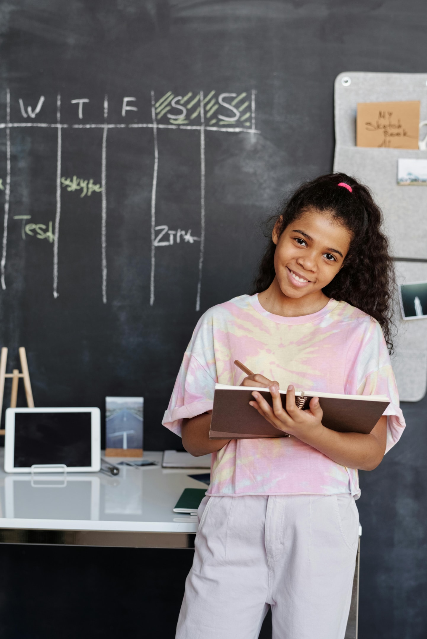 Girl in a pink and white T-shirt standing in front of a blackboard and holding a brown notebook.