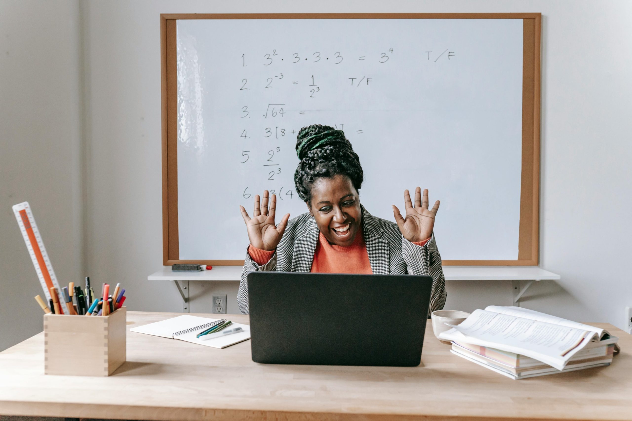 A happy female teacher is using a laptop at a school desk while smiling with her hands in the air. 