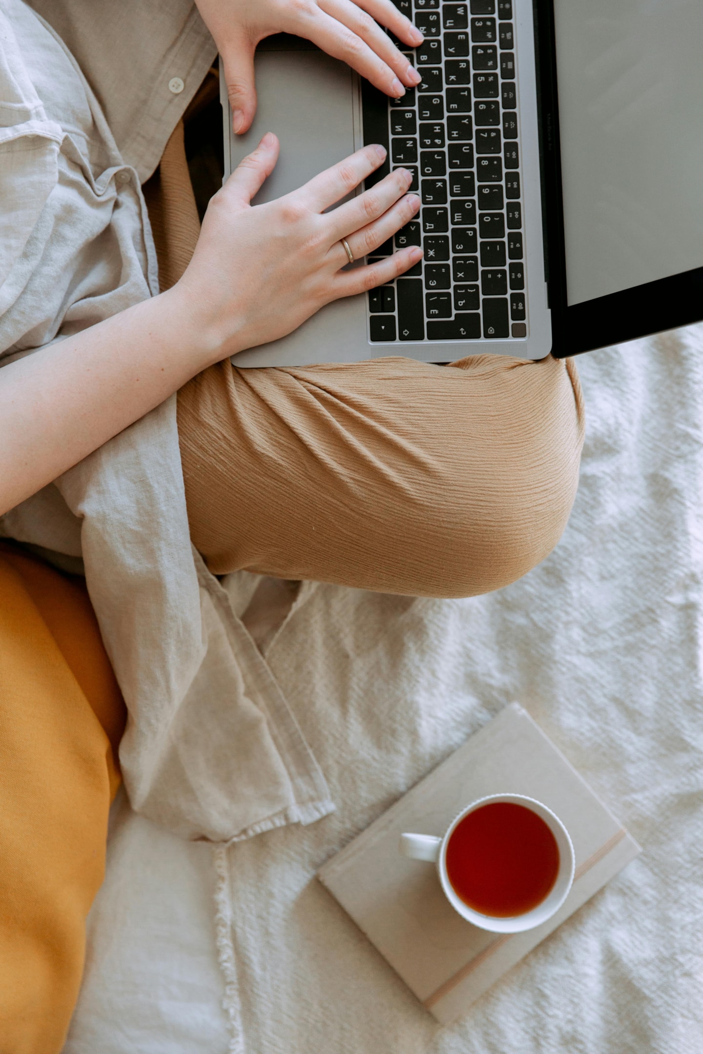 A close up of a woman typing on a laptop with a cup of tea sitting on top of a notebook next to her. 