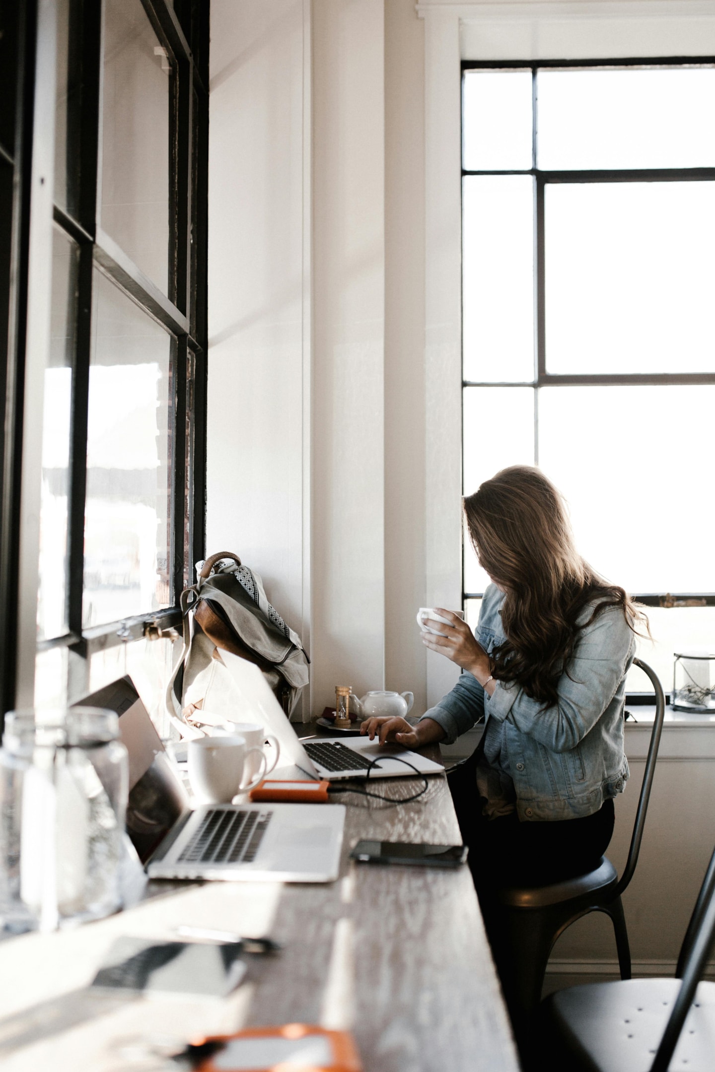 A woman in a gray jacket setting in front of a desk in an office with a coffee in her hand. 