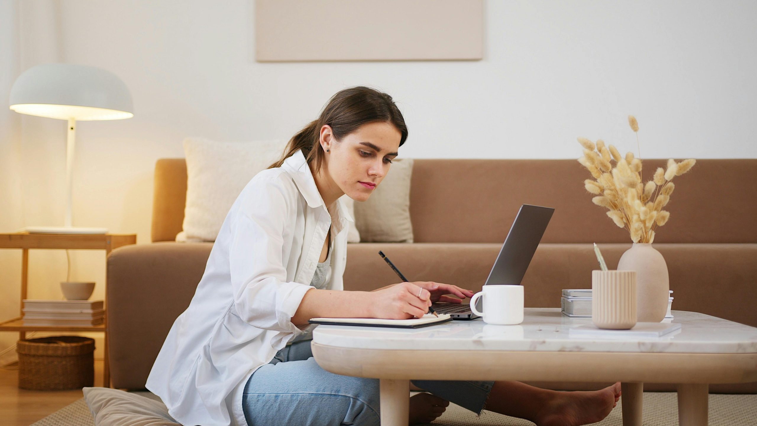 Uma jovem sentada no chão da sala de estar usando um laptop enquanto faz anotações.