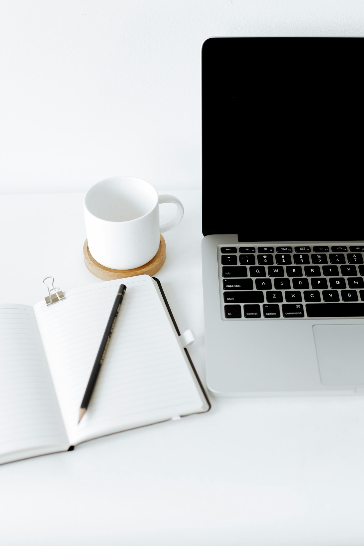 A MacBook, coffee mug and open diary with a black pencil on a white desk.