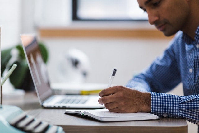 A man writing in a notebook with a laptop in front of him.