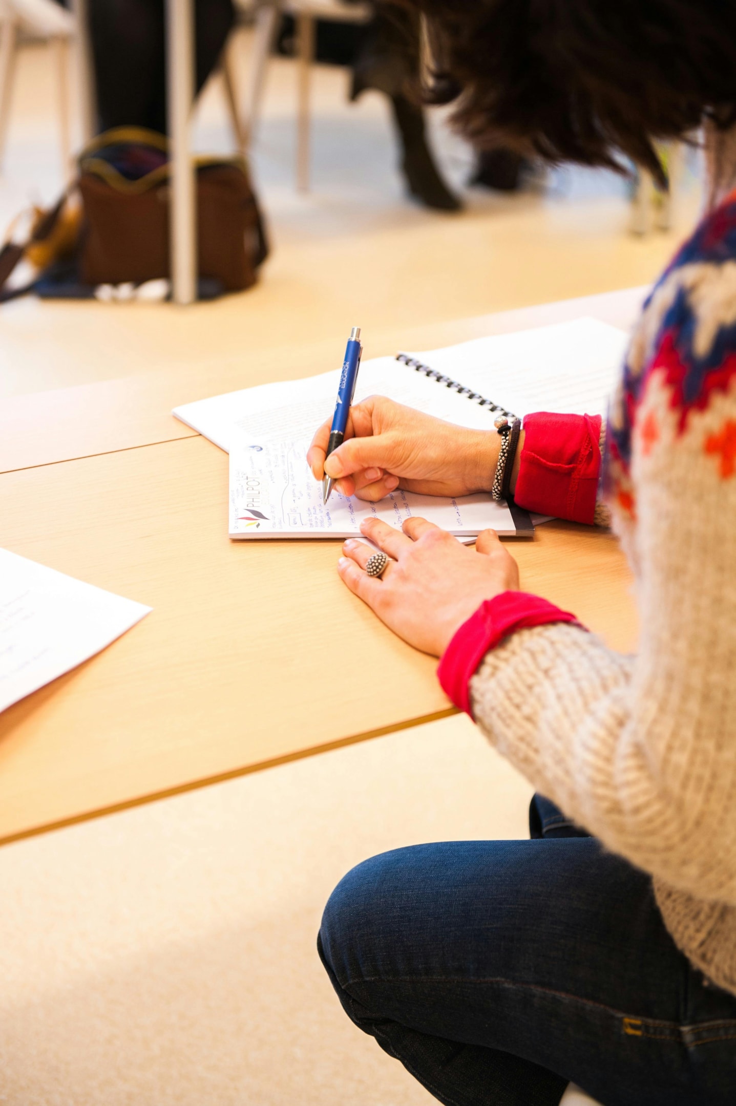 A woman holding a blue pen is sitting at a desk and writing in a notebook. 