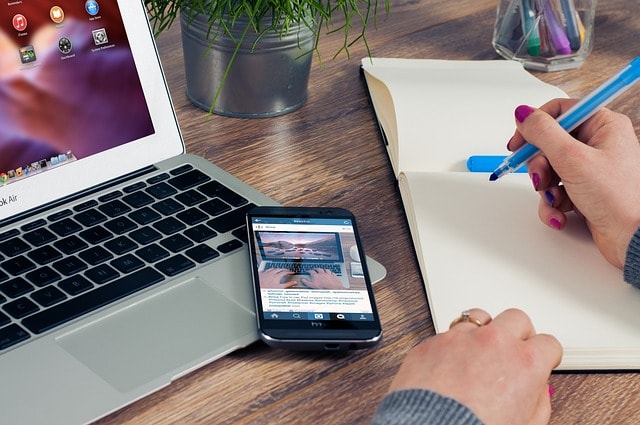 A person writing in a notebook with a blue pen with a laptop and a smartphone next to them.