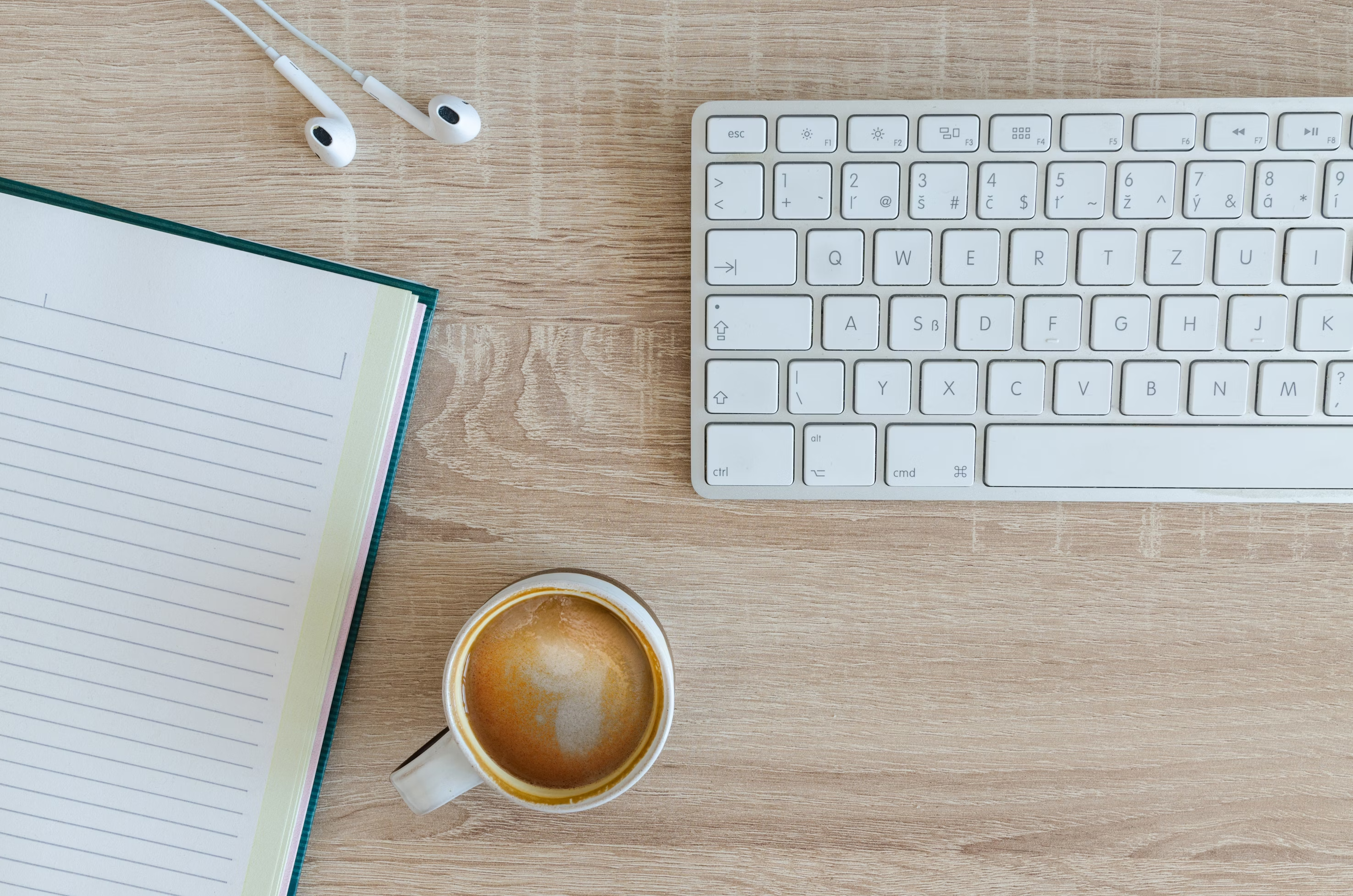 An Apple keyboard, earphones, a notebook, and a cup of coffee on a wooden desk.