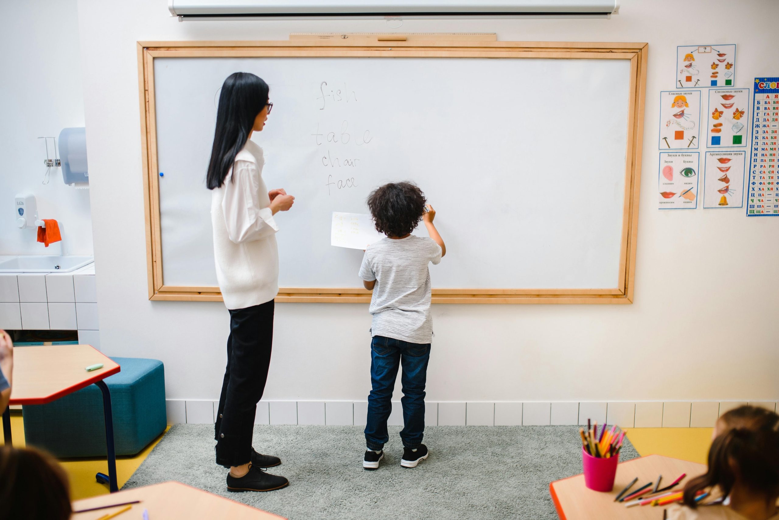 A woman and a boy standing in front of a whiteboard with the boy writing on the board.