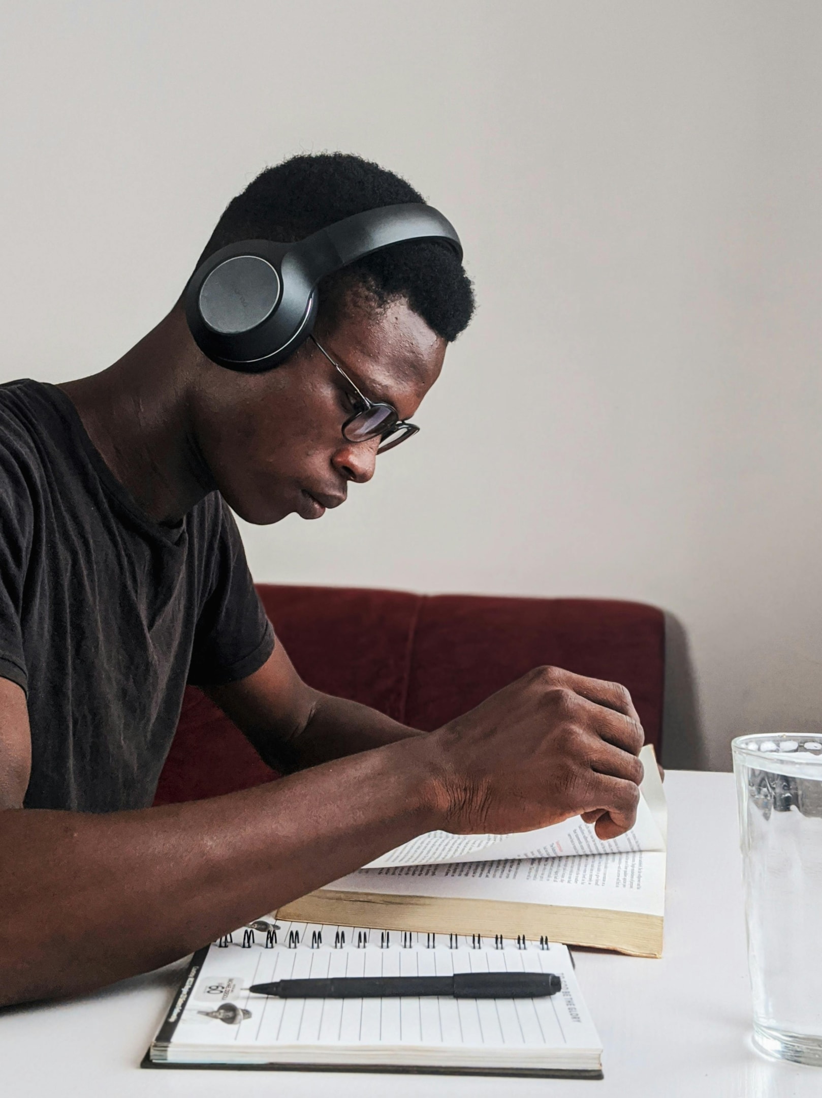 A man wearing a black t-shirt and headphones reading at a desk with a notebook next to him.