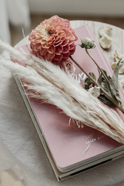 A pink notebook under some pink and white flowers on a white marble desk.