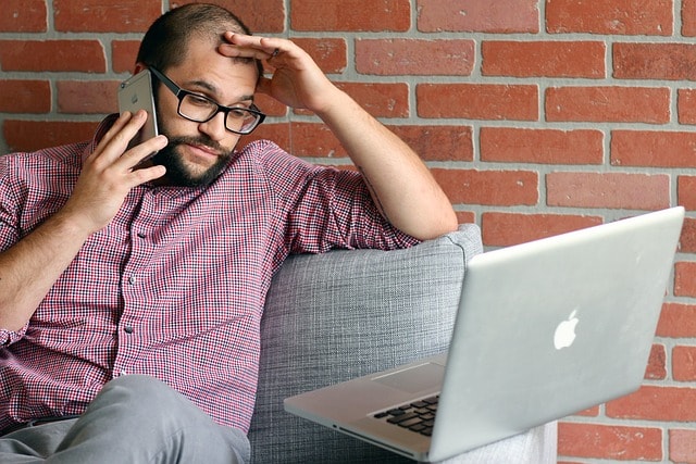 A person holding their head while sitting on a sofa with a laptop and talking on a smartphone.