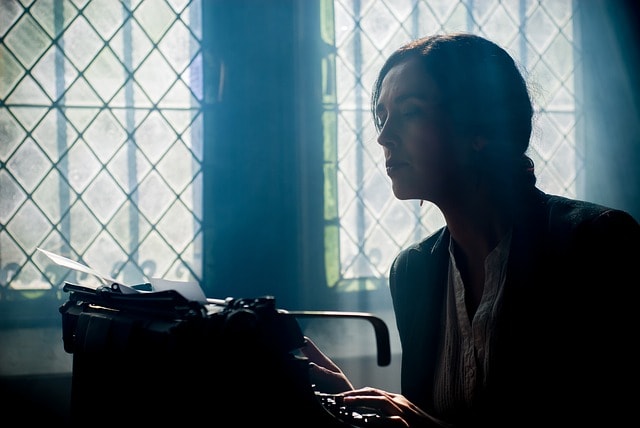 Someone sitting at a desk near a window and typing on a typewriter.