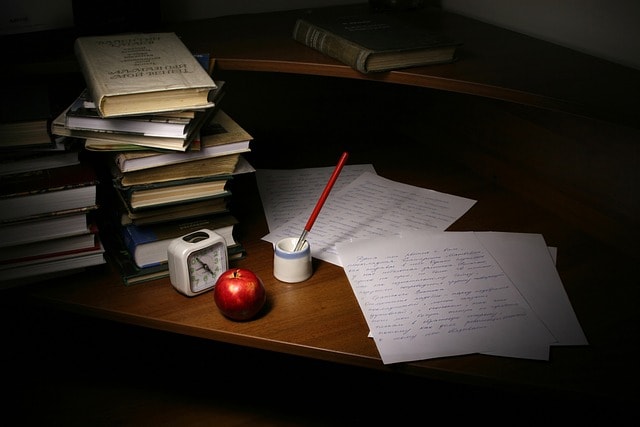 A wooden desk with pieces of paper, books, an apple, a clock, and an inkwell with a pen in it.