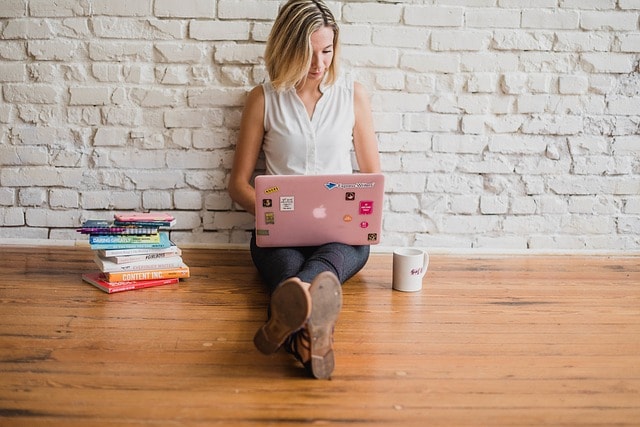 A woman sitting on a wooden floor with her laptop and a stack of books and coffee mug next to her.