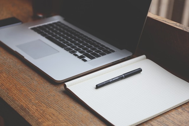 A laptop and notepad with a pen on a wooden desk.