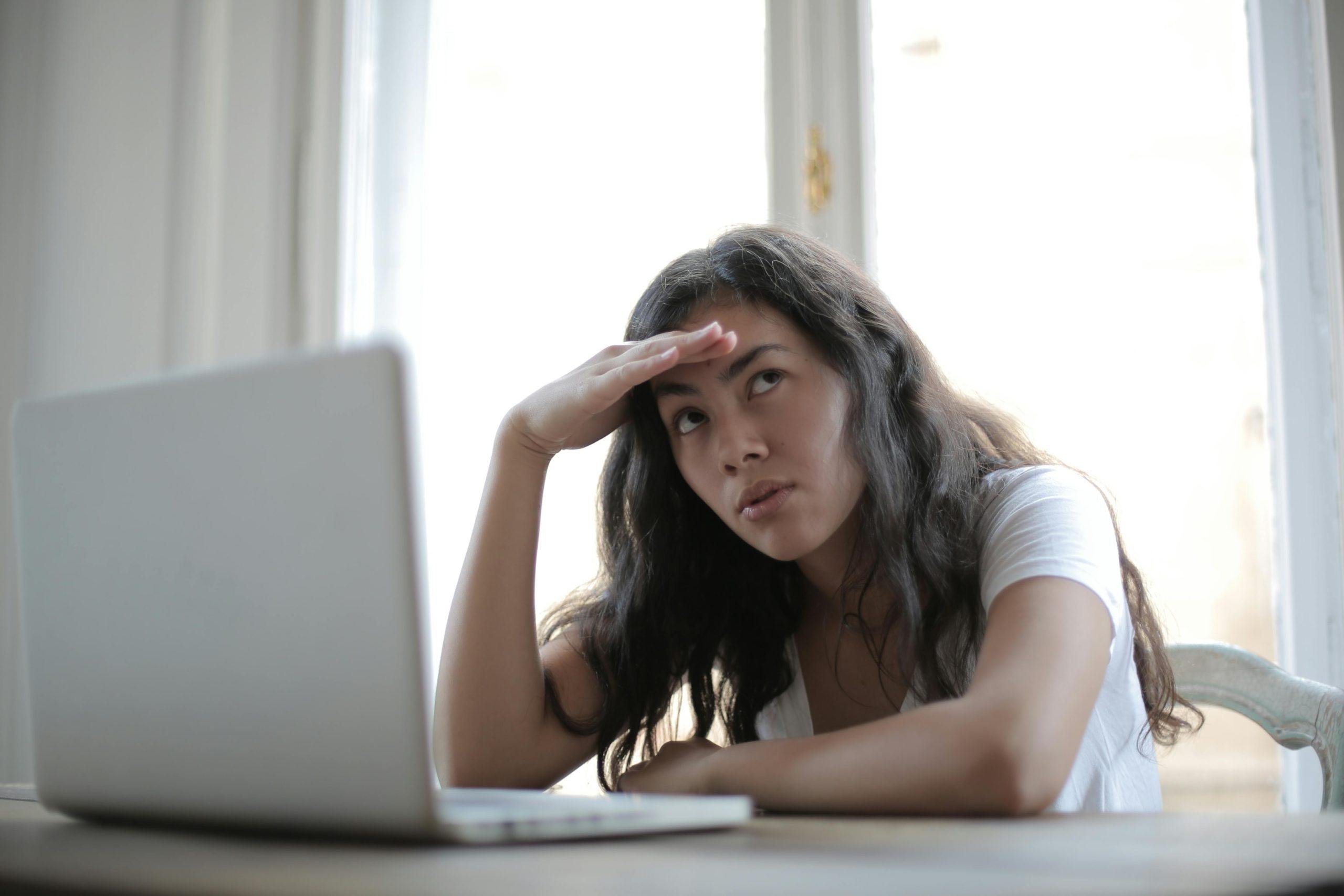 A frustrated looking female student resting her head in her hand while sitting in front of a laptop at a desk.