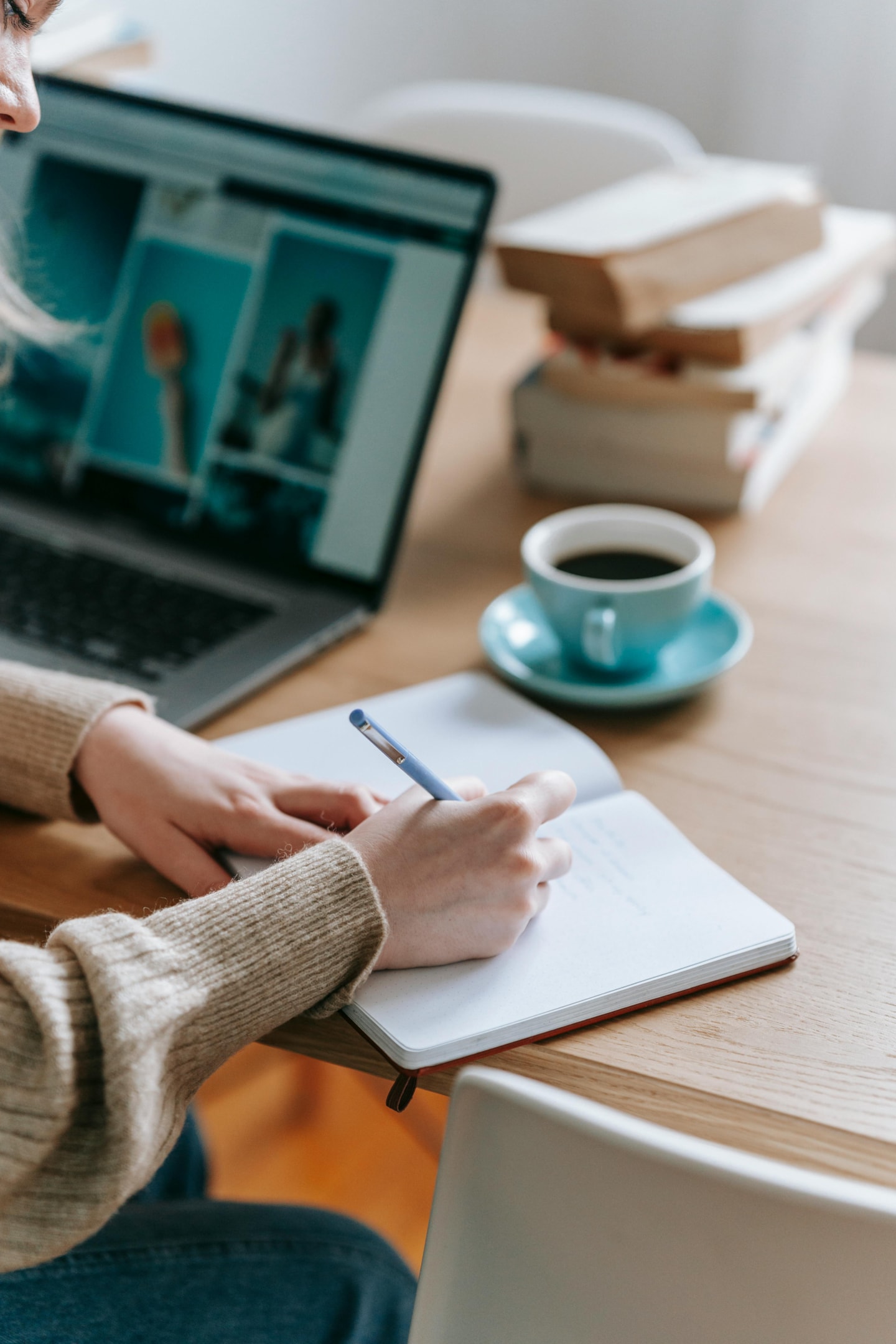 A woman is writing at a desk in a blank notepad with a laptop, books, and a cup of coffee next to her.