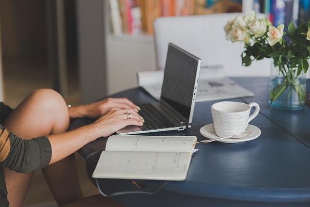 A woman sitting on a desk and typing on her laptop with a cup of coffee and a notebook next to her.