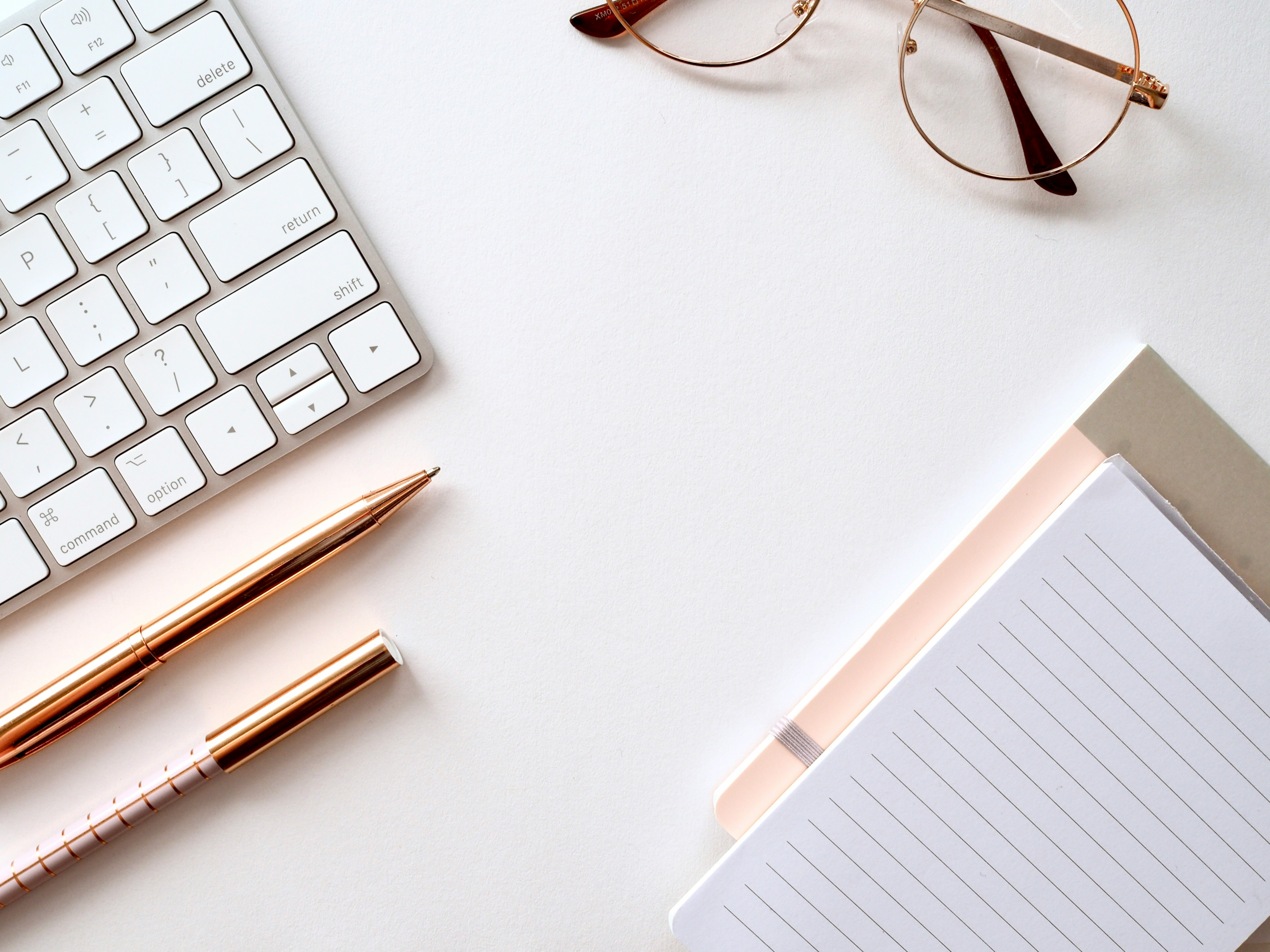 A keyboard, a pen, reading glasses, and a notepad all placed on a white surface.