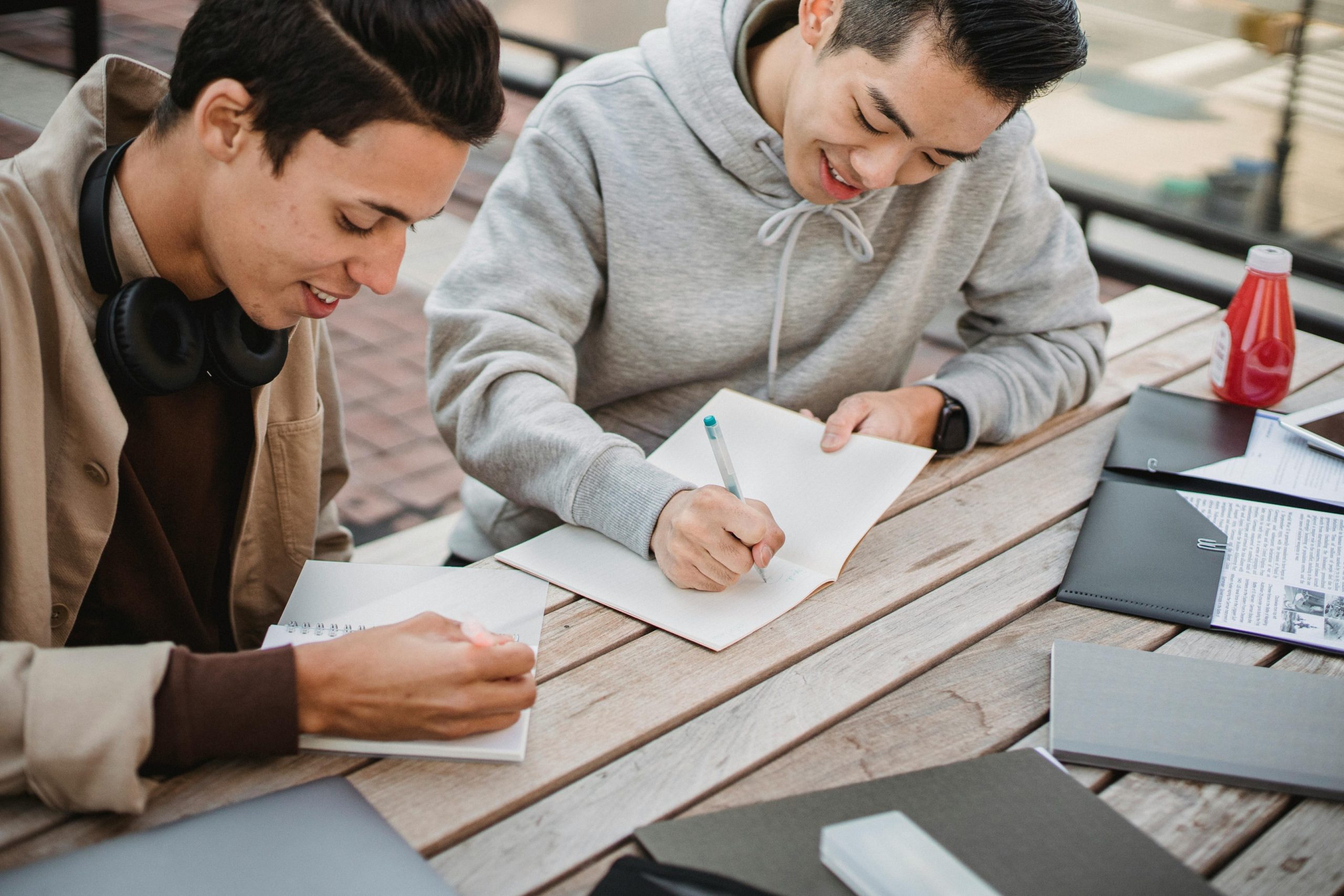 Two male students smiling and writing notes in their notebooks.