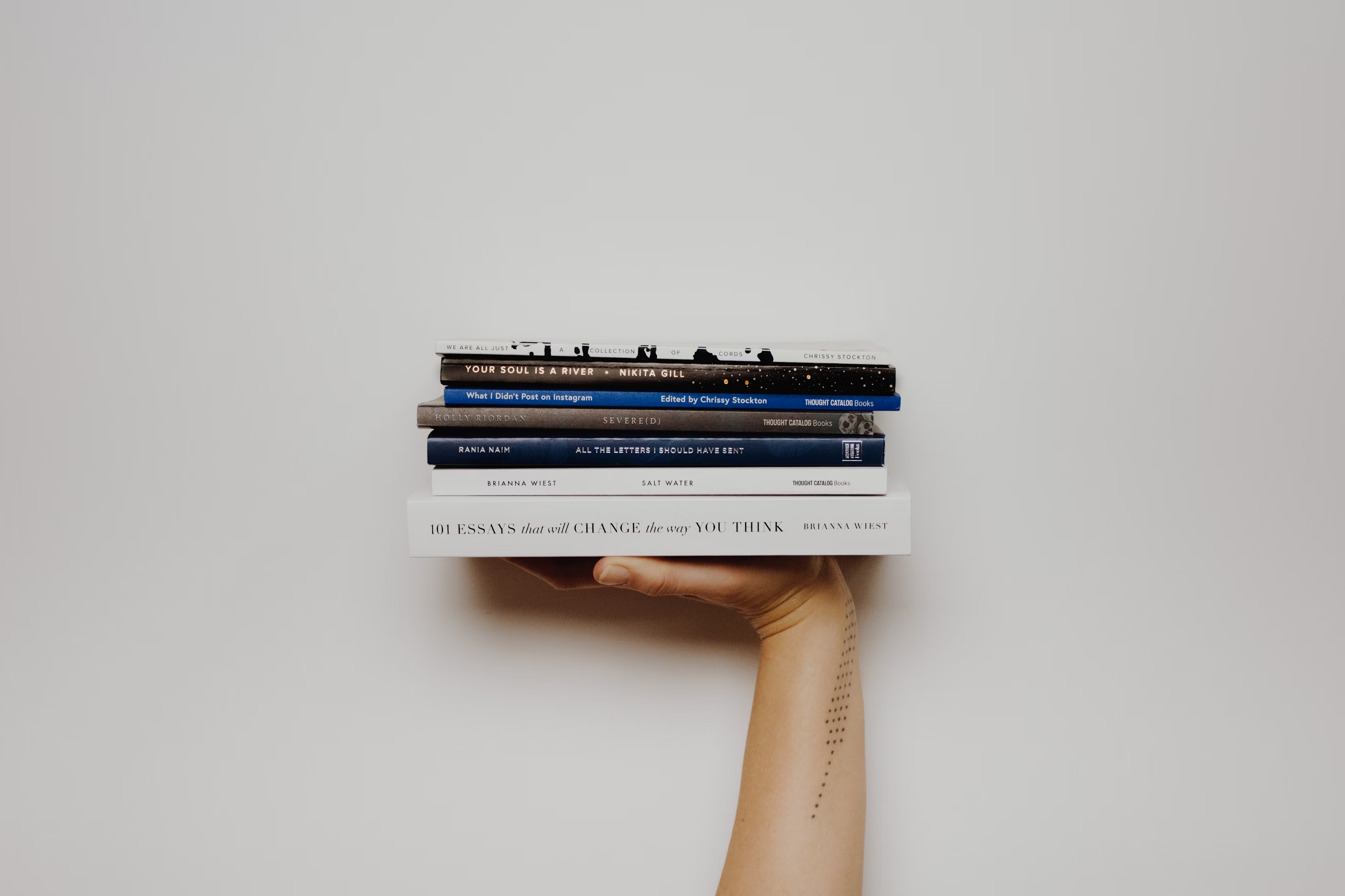 A person holding a pile of books about essays up against a white wall.