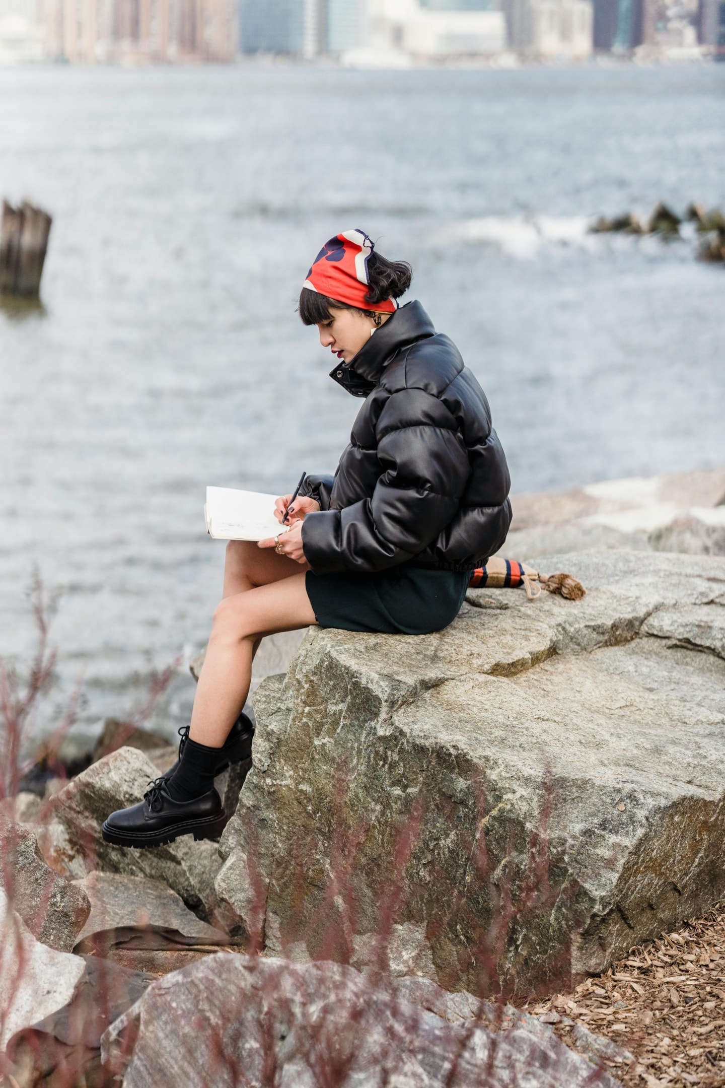 A young woman writing in a notepad while sitting on a rock on the banks of a river.