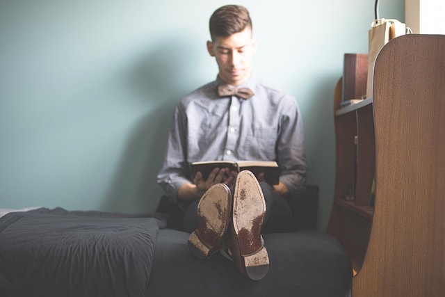 A student with a bow tie sitting on his bed and reading a book.