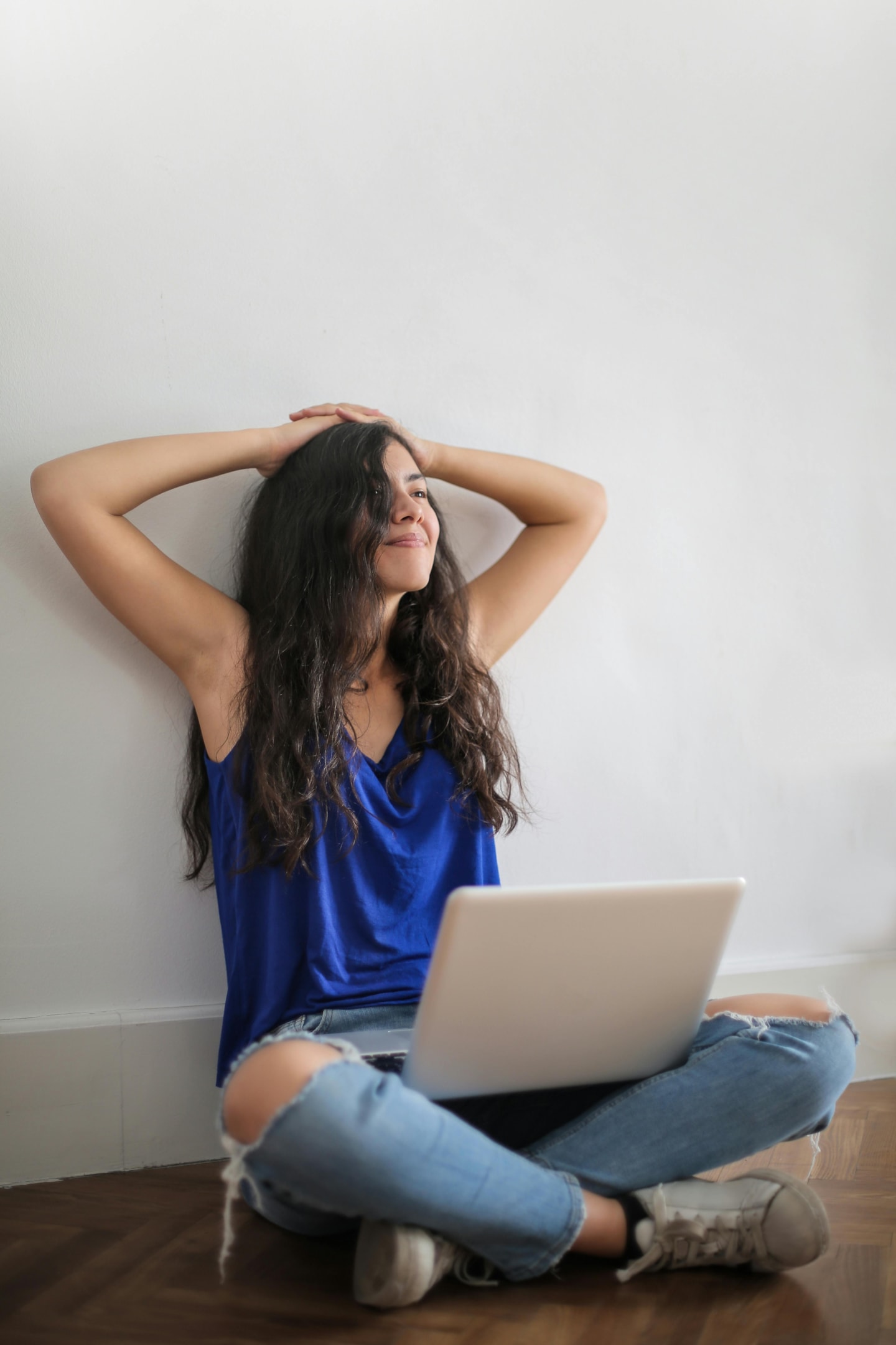 A smiling woman sitting on the floor of a room with a laptop resting in her lap. 