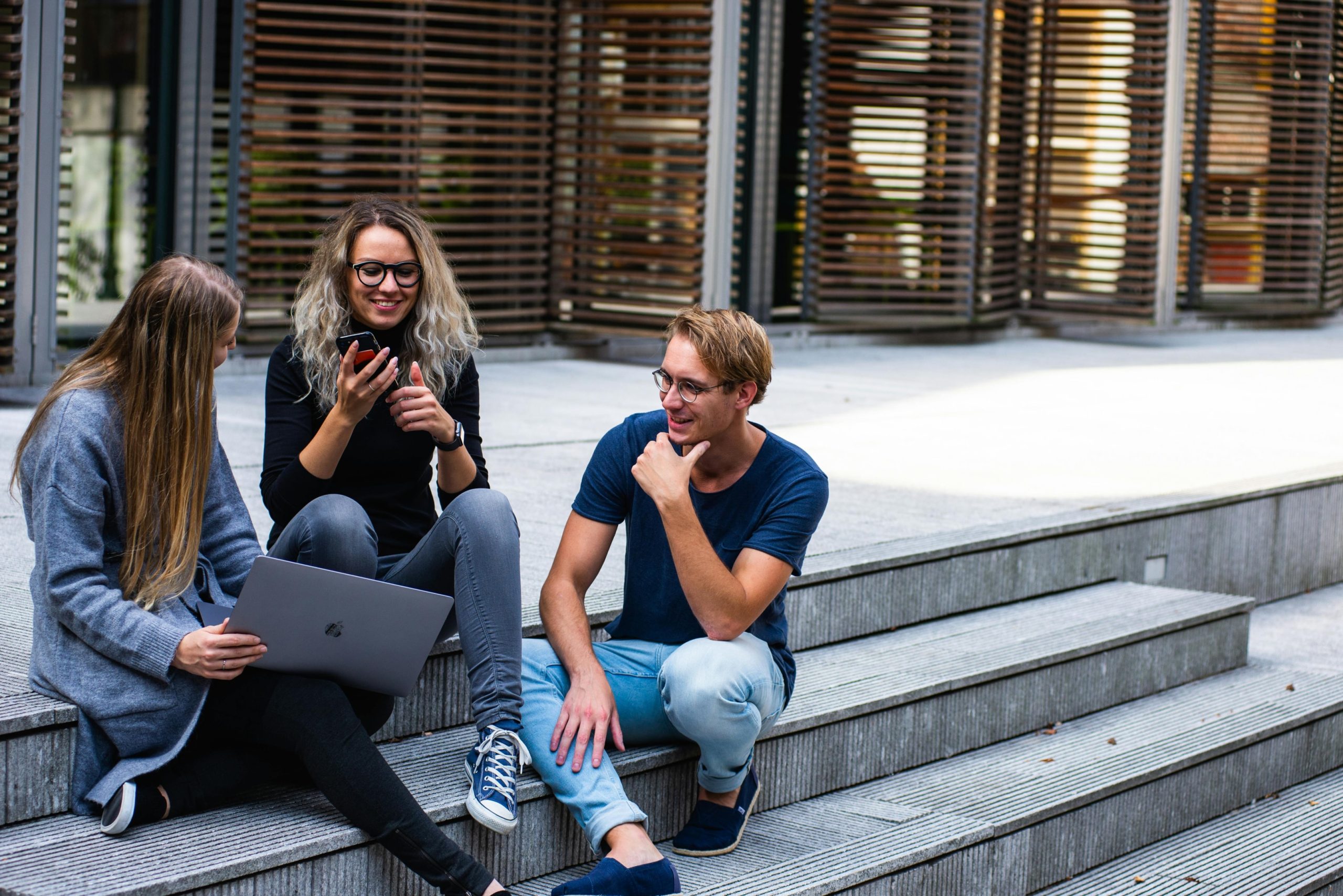 Three students smiling while sitting on college campus stairs outdoors.