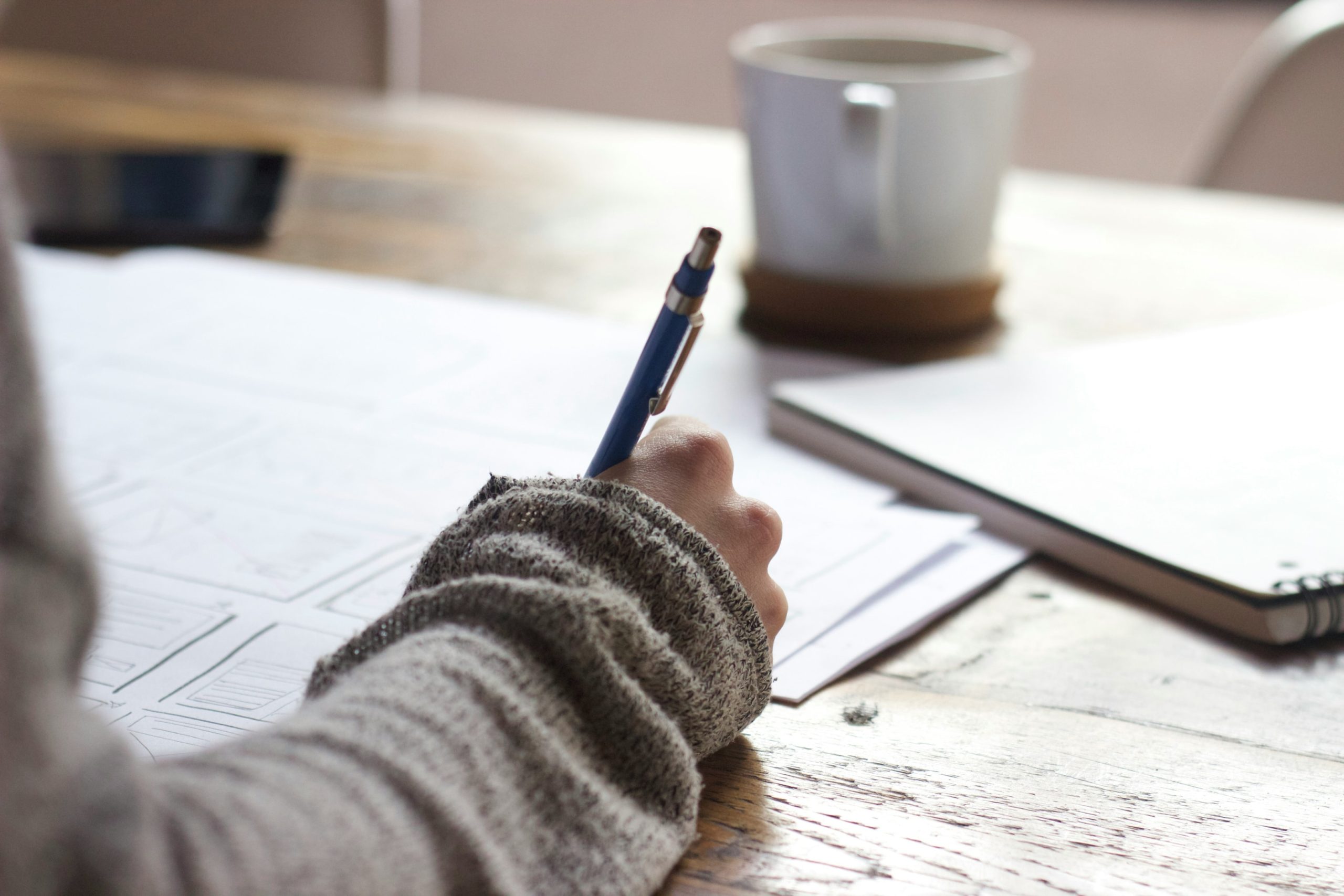 A person writing some notes and surrounded by a notebook and a coffee mug.