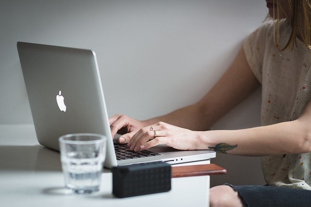 A woman writing on her Macbook with a glass of water next to her.