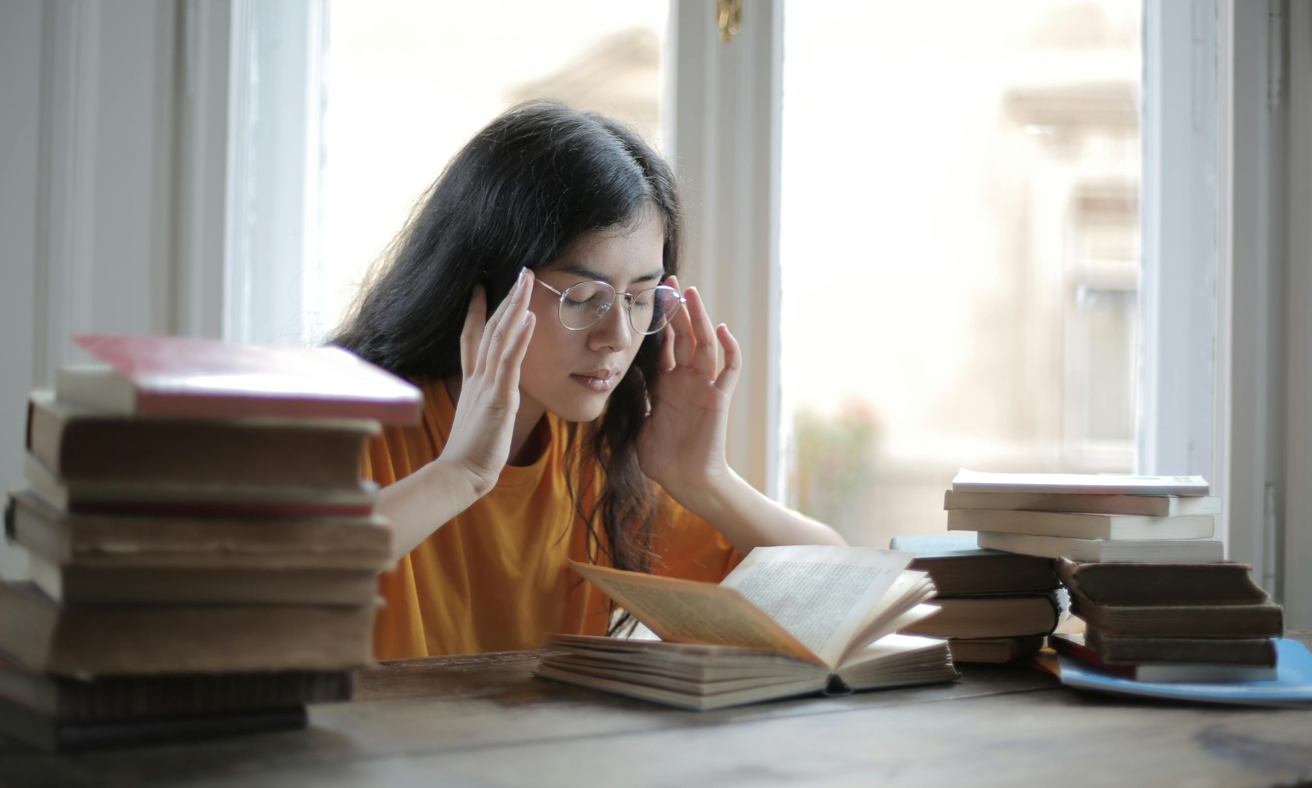 A woman holding her head with her eyes closed while sitting at a desk with piles of books either side of her. 