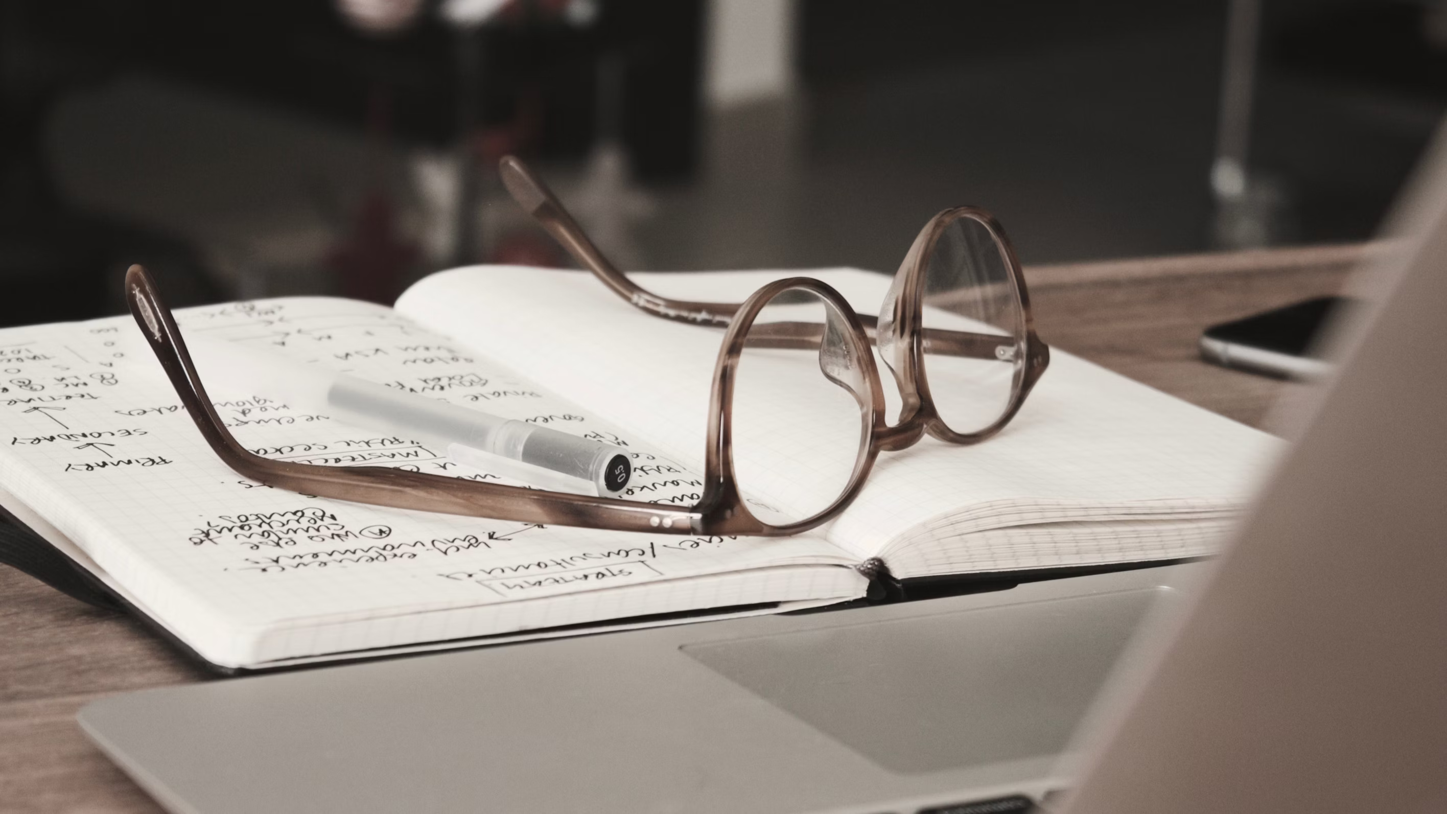 Glasses with gray frames on top of an open notebook, along with a pen.