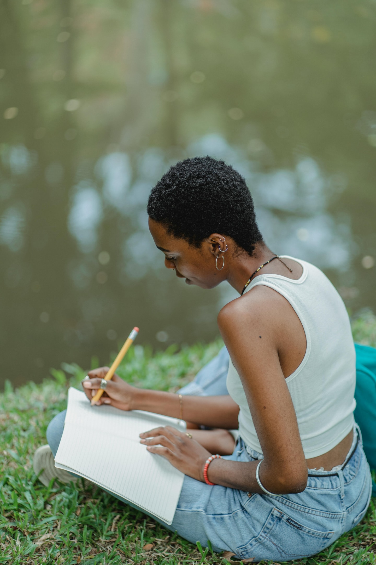 A woman sitting next to a river in a park while writing in a notebook.