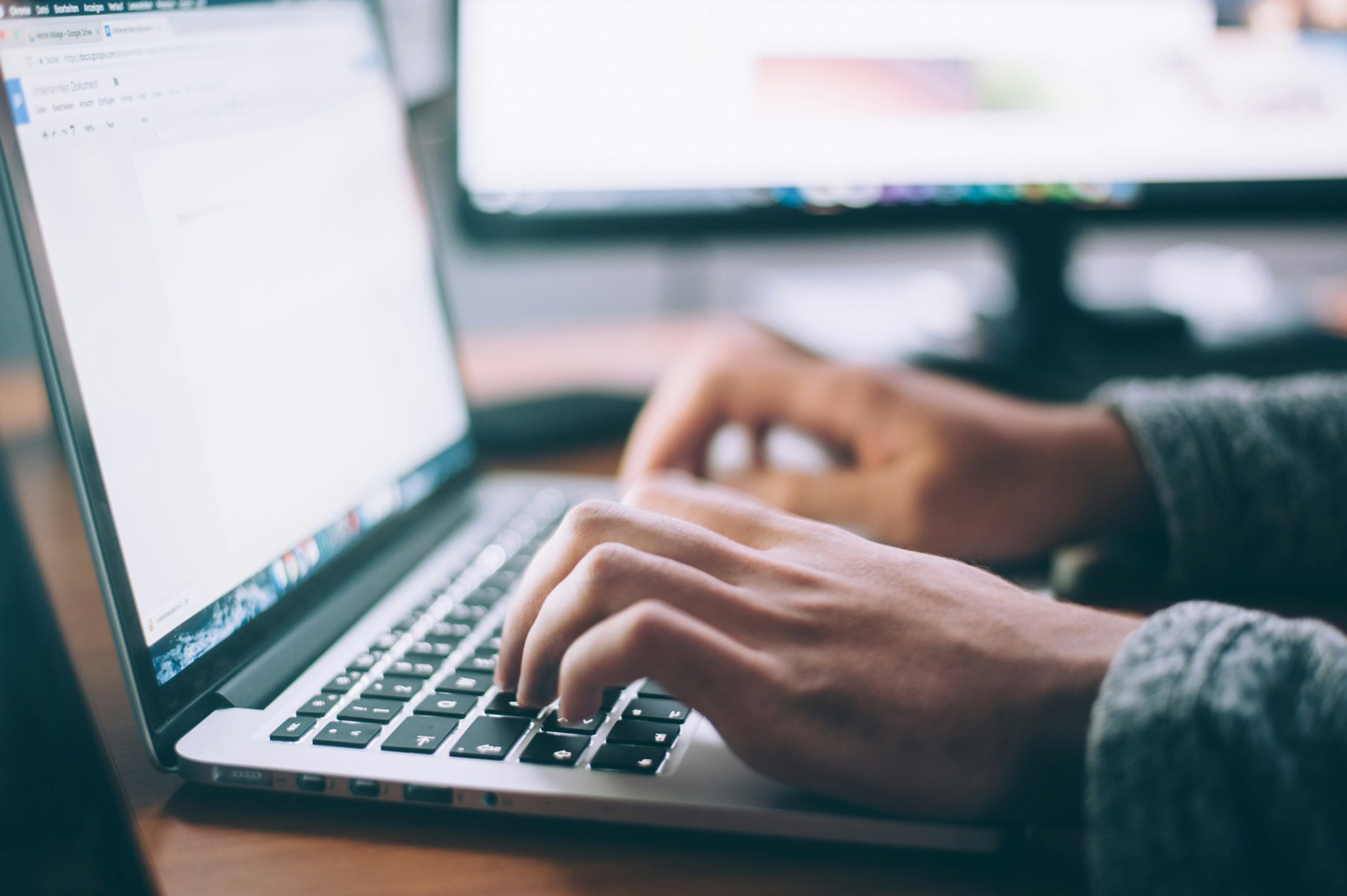 A person typing on a MacBook with another computer screen in the background.