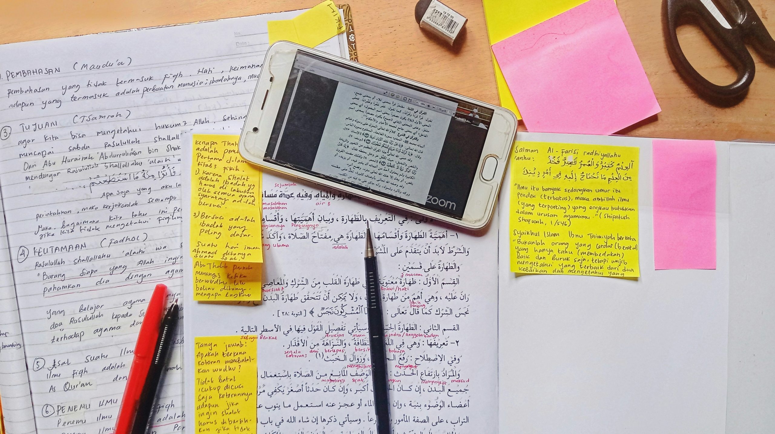 A smartphone, some pink and yellow sticky notes, a red pen, and pieces of paper on a desk.