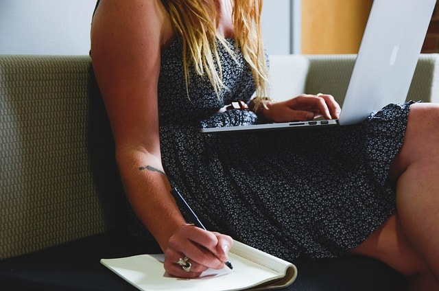 A woman writing on a piece of paper with a laptop on her lap.