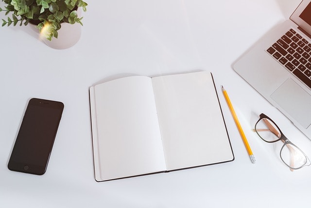 A laptop, a phone, reading glasses, a pencil, and a notebook on a white surface.